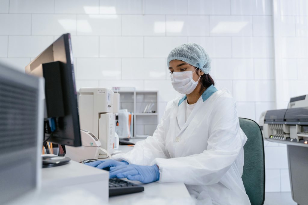 Health and Care Technologies. A scientist in a helth care lab wearing gloves and a mask types at  a computer.